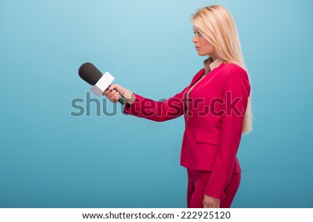Half-length portrait of fair-haired serious TV presenter wearing great red jacket and cream-colored shirt standing aside interviewing someone. Isolated on blue background