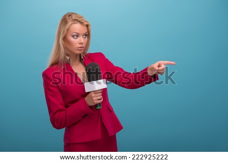 Half-length portrait of very beautiful fair-haired anxious TV presenter wearing great red jacket and cream-colored shirt standing aside speaking into the microphone pointing at someone. Isolated on
