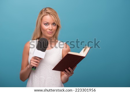 Half-length portrait of lovely fair-haired TV presenter wearing pretty white dress standing to us holding a microphone reading a book. Isolated on blue background
