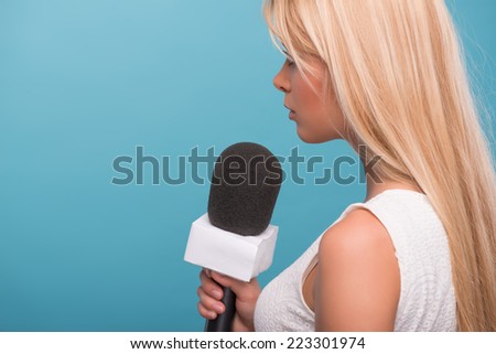 Half-length portrait of lovely fair-haired TV presenter wearing pretty white dress standing back to us holding a microphone. Isolated on blue background