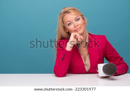 Half-length portrait of lovely fair-haired TV presenter wearing great red jacket and cream-colored shirt sitting at the table holding a microphone dreaming about something wonderful looking at us.