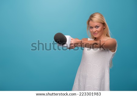Half-length portrait of lovely smiling fair-haired TV presenter wearing pretty white dress holding a microphone wanted to interview everybody. Isolated on blue background