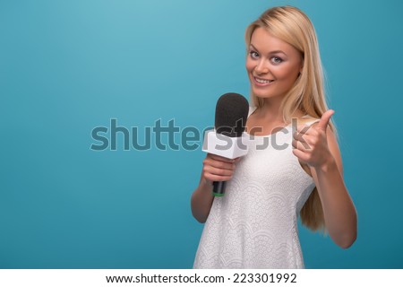 Half-length portrait of lovely smiling fair-haired TV presenter wearing pretty white dress holding a microphone showing that she likes her job. Isolated on blue background