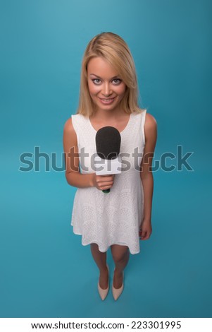 Half-length portrait of lovely smiling fair-haired TV presenter wearing pretty white dress holding a microphone. Top view. Isolated on blue background