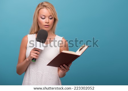 Half-length portrait of lovely fair-haired TV presenter wearing pretty white dress standing to us holding a microphone reading a book. Isolated on blue background
