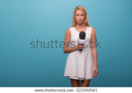 Half-length portrait of lovely fair-haired serious TV presenter wearing pretty white dress standing holding a microphone. Isolated on blue background