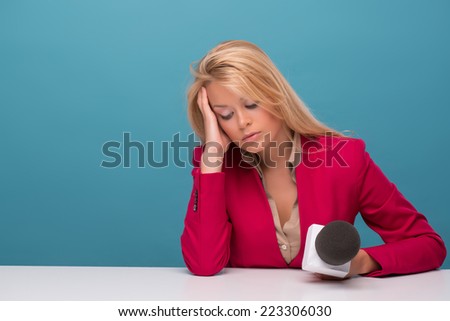 Half-length portrait of lovely fair-haired tired TV presenter wearing great red jacket and cream-colored shirt sitting at the table holding a microphone sleeping at her working place. Isolated on blue