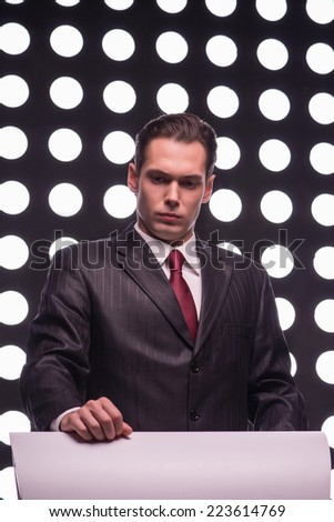 Half- length portrait of handsome TV presenter wearing great black suit and vinous tie standing behind the rostrum thinking hard about something