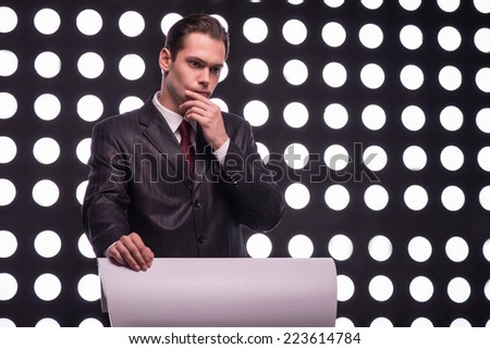 Half- length portrait of handsome TV presenter wearing great black suit and vinous tie standing behind the rostrum thinking hard about something