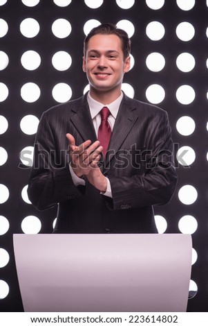 Half- length portrait of young handsome smiling TV presenter wearing great black suit and vinous tie standing behind the rostrum applauding someone