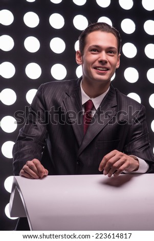 Half- length portrait of young handsome smiling TV presenter wearing great black suit and vinous tie standing behind the rostrum leaning on it listening to someone