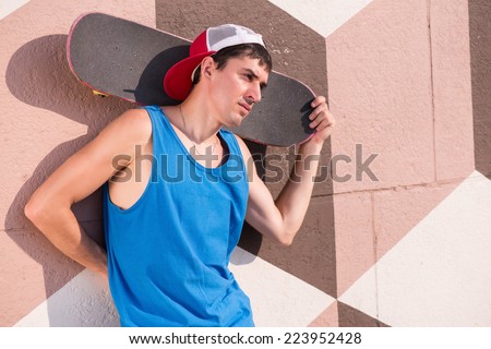 Full-length portrait of young man wearing shorts blue T-shirt and trainers holding the skateboard leaning at the wall