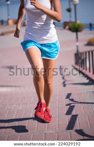 Selective focus on the lovely young woman wearing white T-shirt blue shorts and red jogging shoes running on the quay