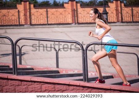 Selective focus on the young sexy smiling woman wearing white T-shirt and blue shorts and red jogging shoes running on the stairs