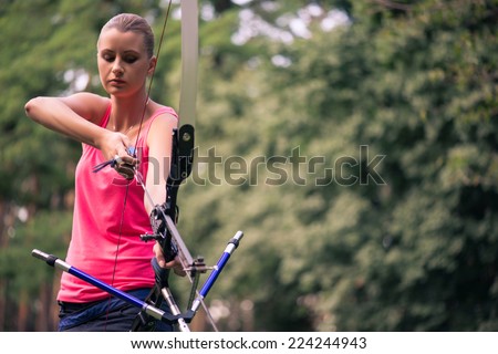Selective focus on the lovely young fair-haired woman standing wearing pink T-shirt and black skirt pulling the bowstring looking for something