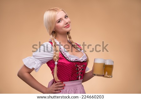 Half-length portrait of young sexy smiling blonde wearing pink dirndl and white blouse standing aside holding in one hand beer mugs. Isolated on dark background