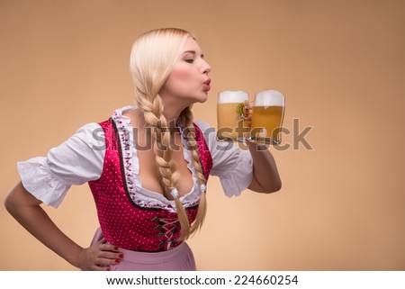 Half-length portrait of young sexy smiling blonde wearing pink dirndl and white blouse standing aside going to drink her favorite beer. Isolated on dark background
