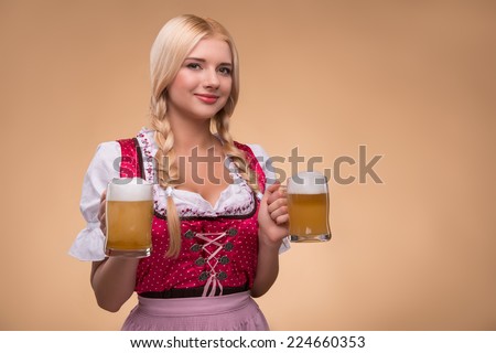 Half-length portrait of young sexy smiling blonde wearing pink dirndl and white blouse holding in both hands beer mugs looking at us. Isolated on dark background