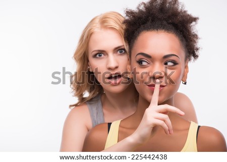 Half-length portrait of young blonde wearing colorful T-shirt hiding behind her smiling friend asking her to be silent looking at us with surprise. Isolated on white background