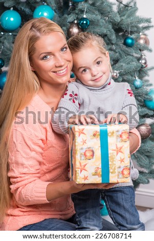 Half-length portrait of beautiful smiling mom and daughter wearing warm sweaters and jeans sitting near the Christmas tree holding nice present