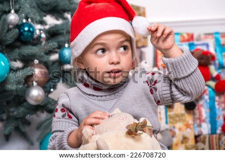 Selective focus on the little cute fair-haired surprised girl sitting on the floor near Christmas presents wearing warm sweater and red cap of Santa Claus playing with the round soft toy