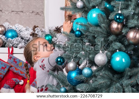 Half-length portrait of the little cute fair-haired girl wearing warm sweater standing aside near the Christmas tree decorating it