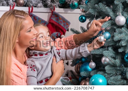 Half-length portrait of the little cute fair-haired smiling girl with her lovely mom wearing warm sweaters and jeans sitting aside near the Christmas tree decorating it