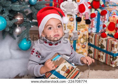 Half-length portrait of the little cute fair-haired girl sitting on the floor near Christmas presents wearing warm sweater and red cap of Santa Claus unpacking her nice present looking at someone