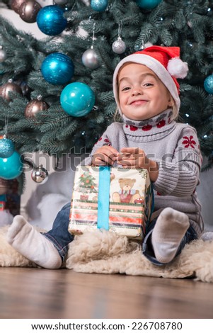 Half-length portrait of the little cute fair-haired smiling girl sitting on the floor near Christmas presents wearing warm sweater and red cap of Santa Claus unpacking her nice present