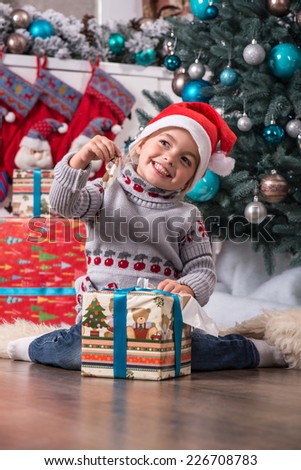 Half-length portrait of the little cute fair-haired smiling girl sitting on the floor near Christmas presents wearing warm sweater and red cap of Santa Claus wanted to unpack her nice present