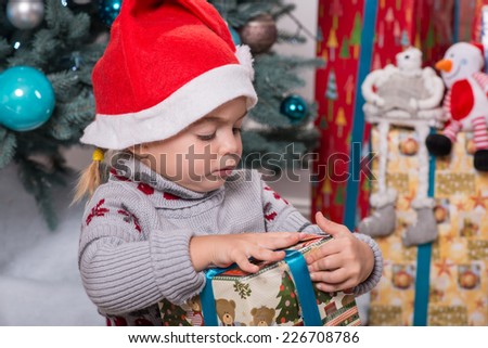 Half-length portrait of the little cute fair-haired girl sitting on the floor near Christmas presents wearing warm sweater and red cap of Santa Claus wanted to unpack her nice present