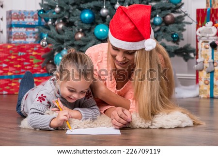Selective focus on the smiling fair-haired mom wearing red cap lying on the floor near the Christmas tree with her little cute daughter writing a letter for Santa Claus