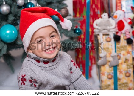 Selective focus on the little cute fair-haired smiling girl sitting on the floor near Christmas presents wearing warm sweater and red cap of Santa Claus thinking about the chocolate