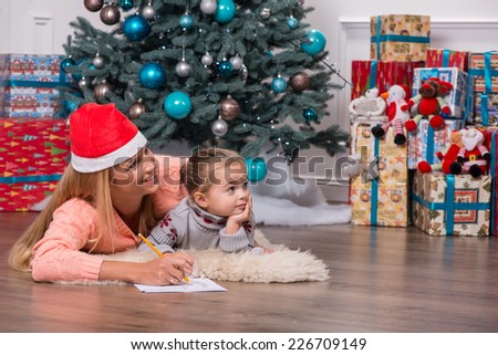 Half-length portrait of smiling fair-haired mom wearing red cap lying on the floor near the Christmas tree with her little cute daughter writing a letter for Santa Claus looking at someone very