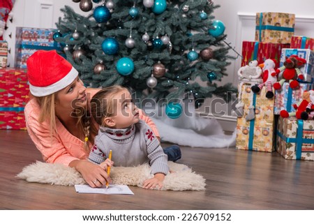 Half-length portrait of smiling fair-haired mom wearing red cap lying on the floor near the Christmas tree with her little cute daughter writing a letter for Santa Claus looking at someone very