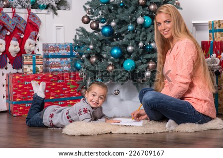 Smiling mom and daughter wearing nice warm sweaters and jeans sitting aside on the floor near the Christmas tree talking and writing a letter for Santa