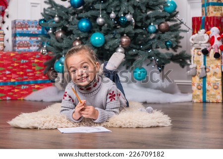 Selective focus on the pretty little fair-haired girl wearing warm sweater and jeans lying on the floor near the Christmas tree with the pencil and paper laughing waiting for Santa Claus