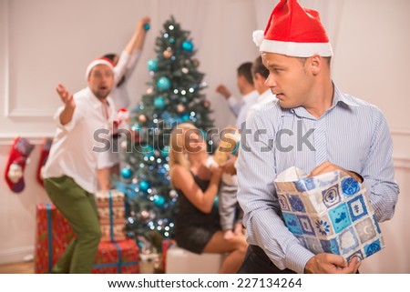 Selective focus on the sly funny guy wearing red cap of Santa Claus standing alone with the present denying giving it back. His friends near the Christmas tree on background