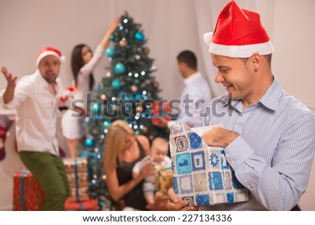 Selective focus on the sly smiling guy wearing red cap of Santa Claus standing alone unpacking the present. His friends near the Christmas tree on background