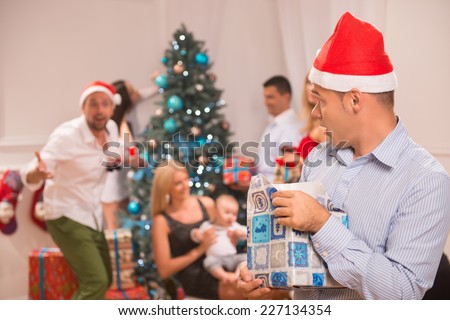 Selective focus on the sly funny guy wearing red cap of Santa Claus standing alone with the present denying giving it back. His friends near the Christmas tree on background