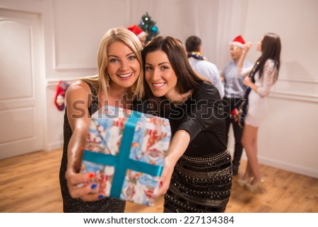 Selective focus on two happy smiling friends standing together showing us their present. Their friends celebrating New Year on background