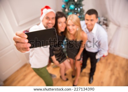 Selective focus on the phone in the hands of smiling guy making photo of his friends standing near the decorated New Year tree on background. Top view