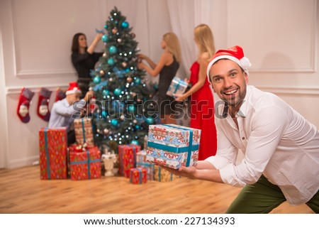 Selective focus on the funny smiling guy wearing red cap of Santa Claus looking out showing us wonderful present. Three beautiful girls wearing fancy clothes and one handsome man decorating the New