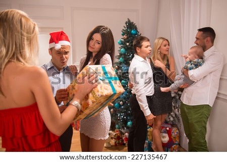 Half-length portrait of the company of happy smartly-dressed people standing in the big light cozy room with the nice New Year tree in the corner greeting each other with the holiday giving trimming