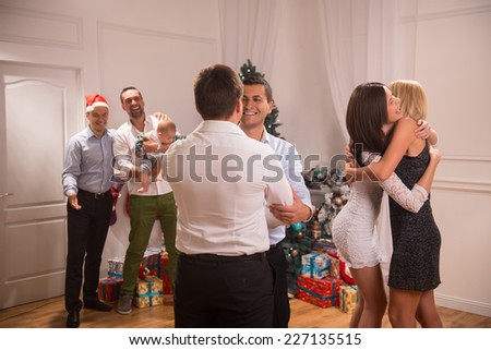 Half-length portrait of the company of happy smartly-dressed people standing in the big light cozy room with the nice New Year tree in the corner greeting each other with the holoday