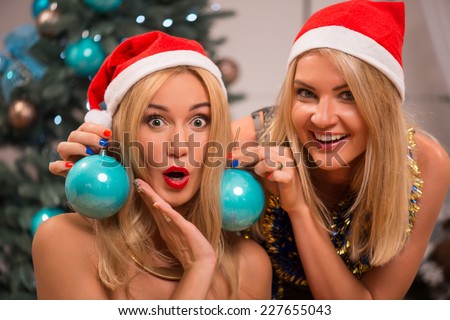 Selective focus on the smiling pretty blonde trying on two turquoise Christmas decorations like earrings at her impressed friend wearing red cap of Santa Claus