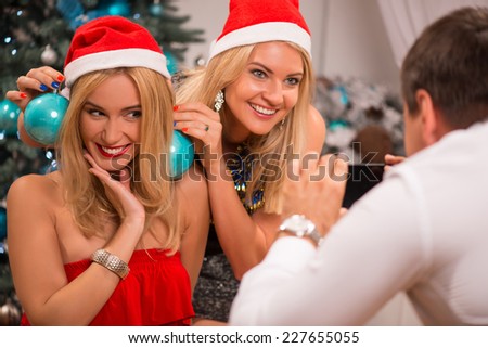 Selective focus on the smiling pretty blonde trying on two turquoise Christmas decorations like earrings at her impressed friend wearing red cap of Santa Claus posing before the man making their photo