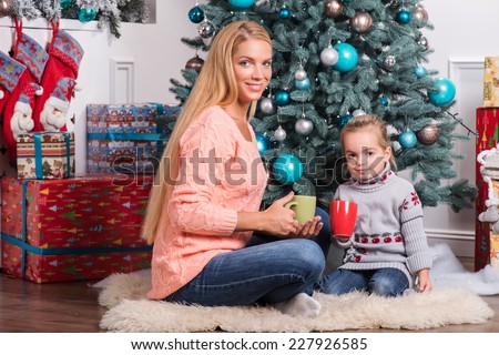 Full-length portrait of smiling beautiful mom and daughter wearing warm sweaters and jeans sitting aside near the Christmas tree drinking delicious cacao waiting for daddy