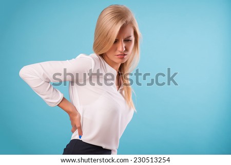 Half-length portrait of beautiful tired blonde wearing white classic blouse and black skirt standing aside having a terrible back pain. Isolated on blue background
