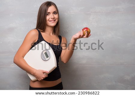 Half-length portrait of lovely smiling dark -haired young woman wearing black vest standing aside holding in one hand very delicious ripe apple and in another electronic balance